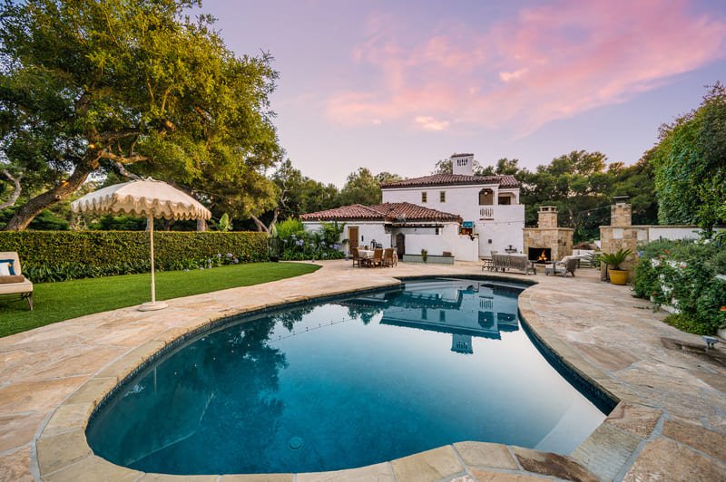 Patio trasero de lujo con piscina iluminada, zona de fogata y comedor exterior bajo un cielo al atardecer.