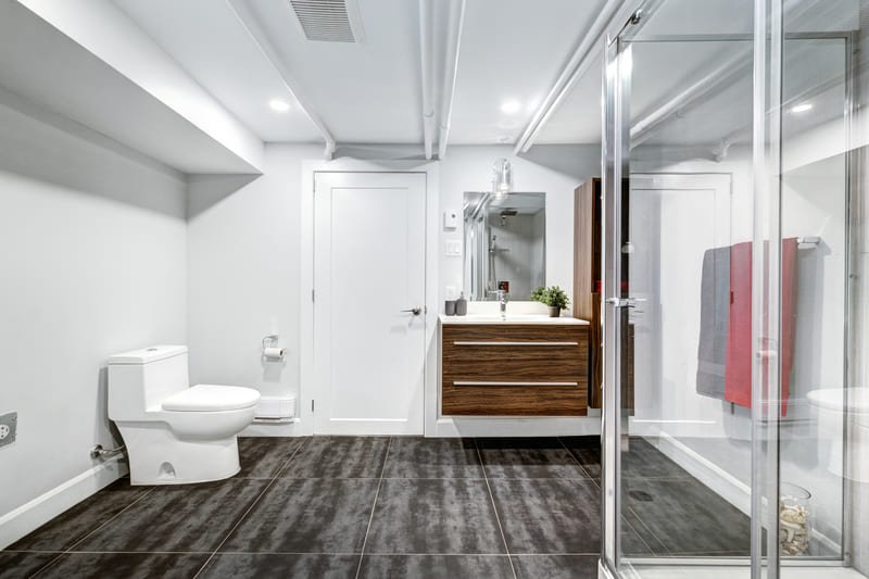 Cuarto de baño amplio con ducha de cristal, piso de loseta oscura, sanitario blanco y mueble de lavabo en madera - Casa Velaris
