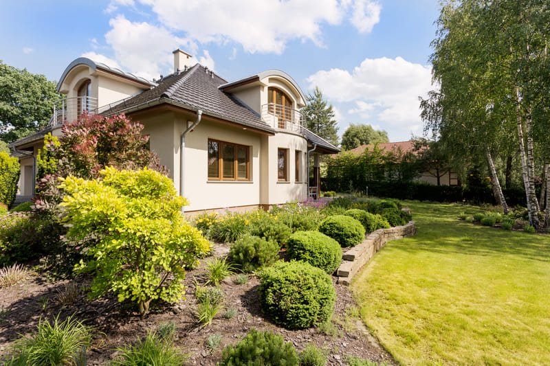 Fachada de casa moderna de dos niveles con jardín paisajista, césped verde y árboles.