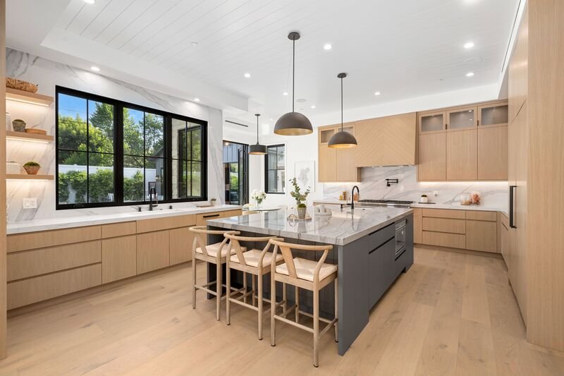 Cocina moderna de concepto abierto con isla central gris, encimeras de mármol y gabinetes de madera clara.