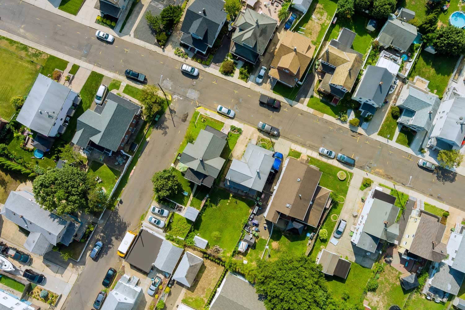 Vista aérea cenital de un vecindario residencial con casas, jardines verdes, calles trazadas y autos estacionados.