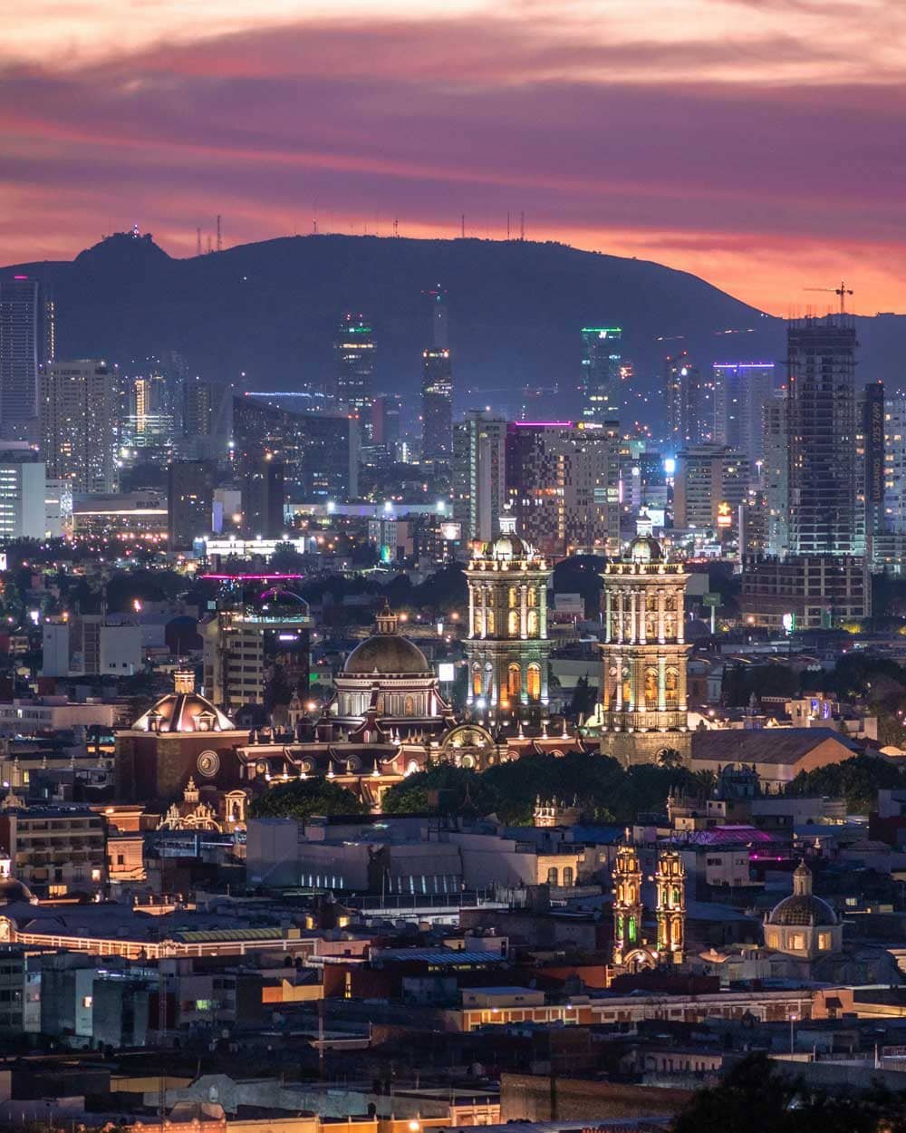Panorámica nocturna de Puebla, México, mostrando la Catedral iluminada y rascacielos modernos al atardecer.